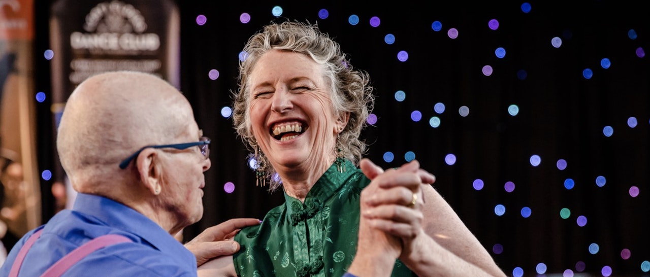 Two people dancing holding hands and other hand on woman's shoulder. Man is on left, bald with glasses wearing a blue shirt with pink braces. The woman is middle aged with curly short slightly grey hair, laughing. In the background are coloured dots