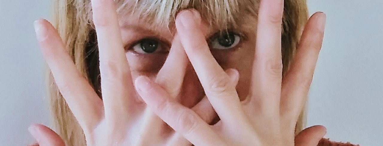 Woman's face hidden behind her fingers of both hands held in front. Woman has straight blonde hair.