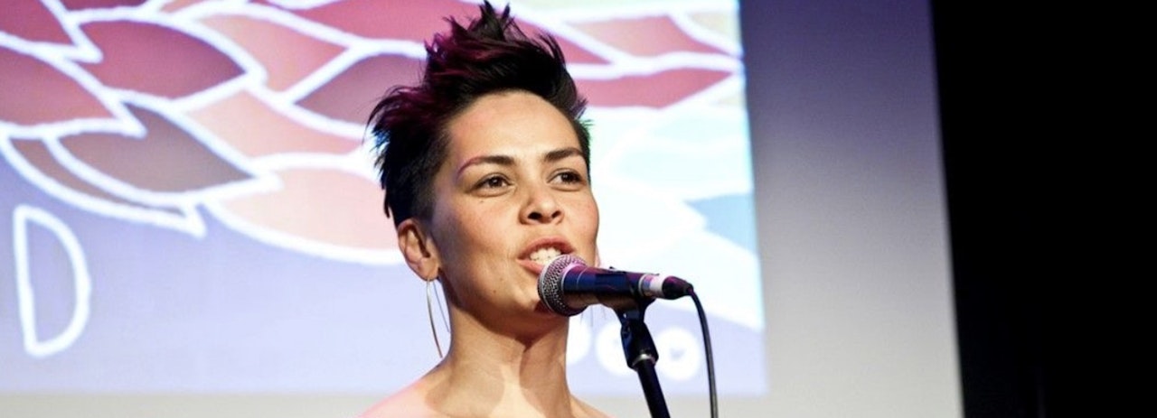 Photograph of woman on stage, from her shoulders up, talking into microphone. Image of leaves projected onto background