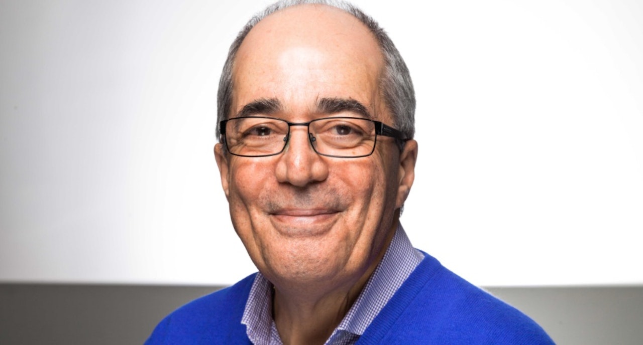 Photograph of a smiling older man wearing eye glasses wearing a shirt and jumper.