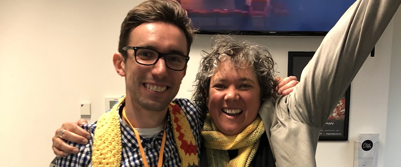 Man and woman with arms around each other. Man has short cropped brown hair and wearing black rimmed glasses. Woman has curly grey hair. Both are smiling and wearing yellow scarves. TV screen in background next to poster on the wall