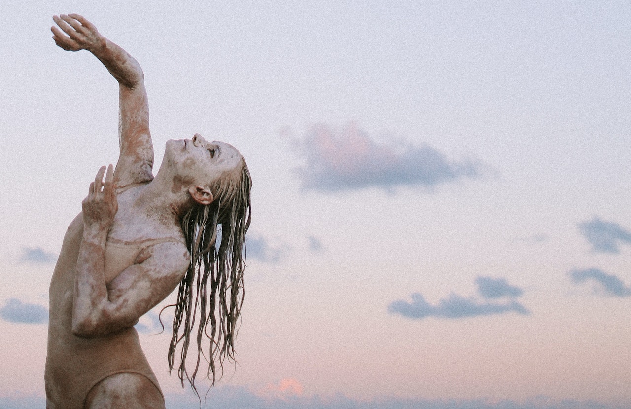 Art photo of Woman covered in mud wearing leotard. Standing in dance position with one arm in the air and the other arm on her chest, looking up. Clouds and sunset in background