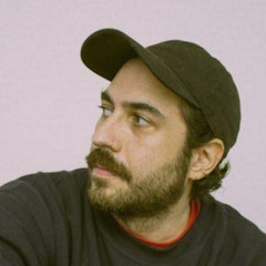 Photo of young man with a beard and wearing a cap and jumper.  Looking off to the side with no face expression