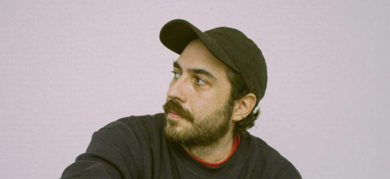 Photo of young man with a beard and wearing a cap and jumper. Looking off to the side with no face expression