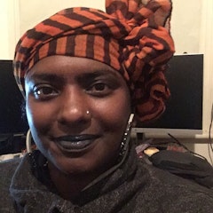 Photo attached description; woman smiling at the camera.  She has brown skin, wears a dark top and large hoop earrings; and an orange striped scarf is wrapped around her head.