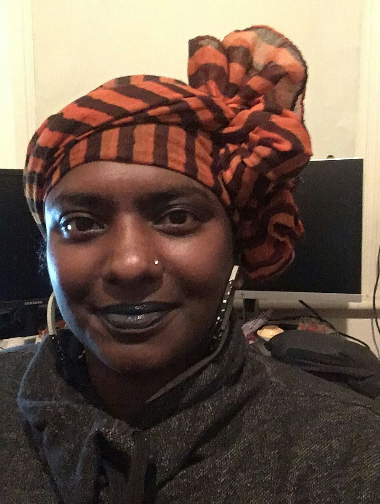 Photo attached description; woman smiling at the camera. She has brown skin, wears a dark top and large hoop earrings; and an orange striped scarf is wrapped around her head.