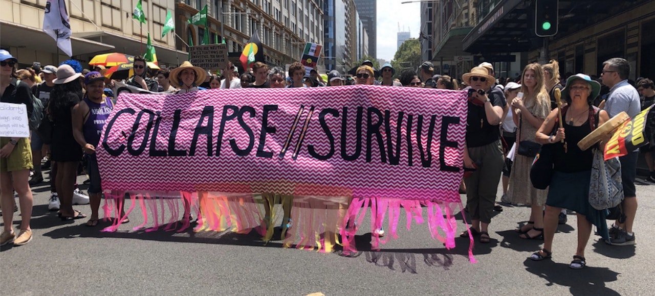 Crowd of people, men and women on a city street. Standing behind a pink banner with black text saying "Collapse//Survive"